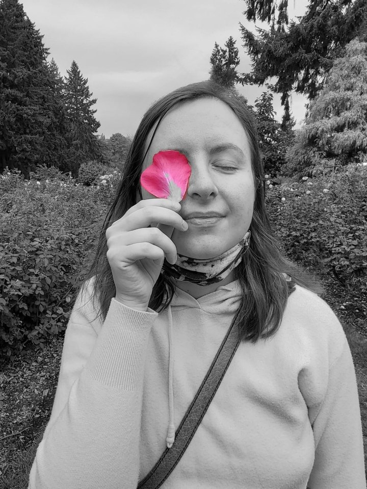 Maria Mishurenko holding a rose petal over one eye in a rose garden.